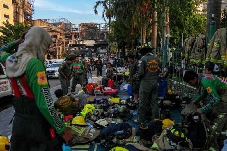 People wearing firefighting gear gather in front of a burned-out building.